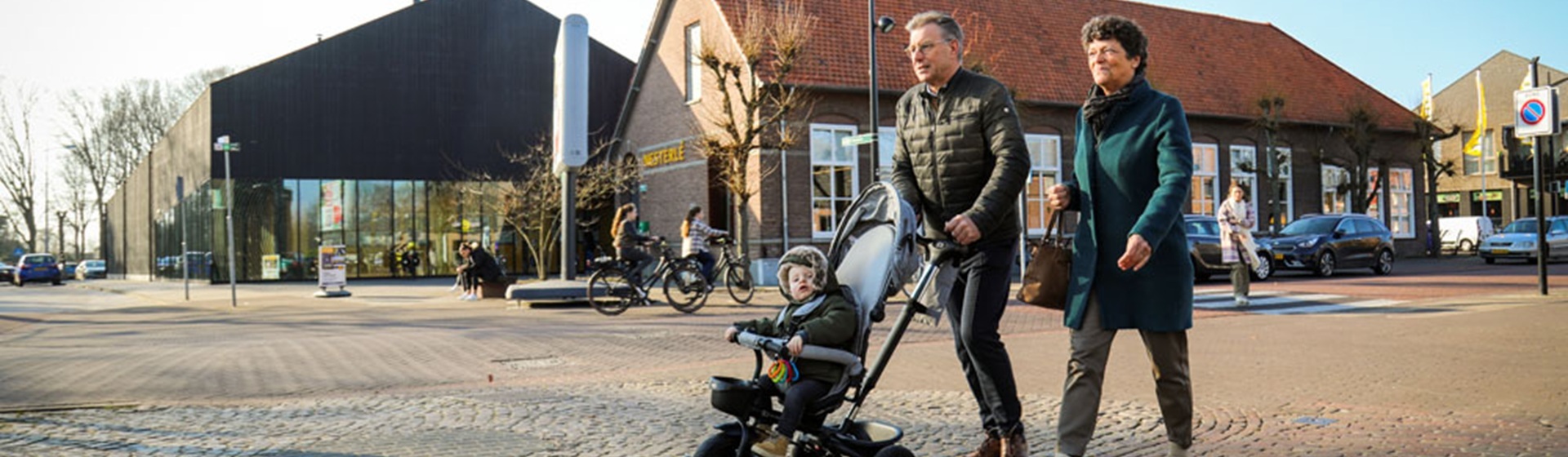 Grootouders wandelen met kinderwagen en kind over straat voor een aantal winkels in Nistelrode